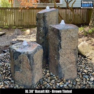 May include: XL 38" Basalt Kit - Brown Tinted. A three-piece outdoor water fountain with a natural, textured appearance. Each piece is a different height, with water spouts at the top. The fountain is surrounded by small, dark-colored stones.