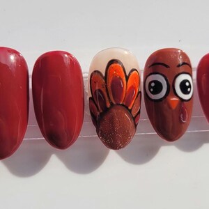 May include: A set of five artificial nails. Two nails are a solid, glossy, dark red. The middle nail features a turkey design with a brown body, orange and brown feathers, and a glittery base. The fourth nail is a turkey face, and the last nail is a solid, glossy, dark red.