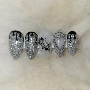 May include: Five silver glitter oval-shaped fake nails with black drip designs. One nail has a spider web design, one has a spider, and one has a heart.
