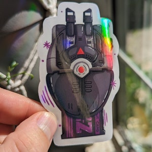 May include: A holographic sticker of a black robot hand with a red eye and a purple ZUT CARD label. The sticker has a rainbow iridescent effect.