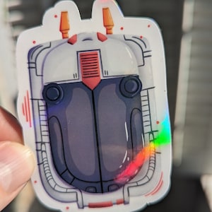 May include: A holographic sticker of a cartoon robot head with a white, gray, and red color scheme. The robot has two orange antennas on top of its head and two black eyes.
