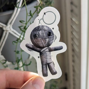 May include: A gray and white yarn doll with a key chain attached to its head. The doll has a simple face with two black eyes and a small mouth. The doll is wearing a gray and white yarn outfit.