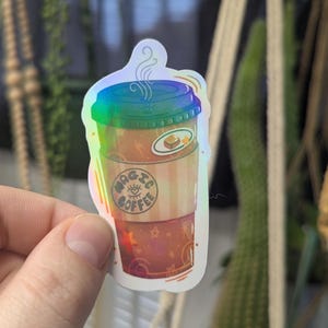 May include: Holographic sticker of a coffee cup with the words "Magic Coffee" and an eye symbol. The cup has a blue and green lid, a brown body, and a red bottom with star details. A small, round sticker with a pastry is on the cup.