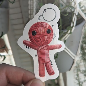 May include: Red yarn doll keychain with a black star and a silver key ring. The doll has black eyes and a red yarn body.