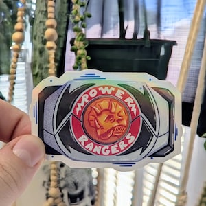 May include: A holographic sticker featuring the Power Rangers logo. The design includes a red and gold dinosaur head emblem, surrounded by the words "POWER RANGERS" in white lettering. The sticker has a black and silver border.