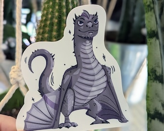 May include: A sticker featuring a cartoon illustration of a purple and grey dragon with wings, standing on a white background.