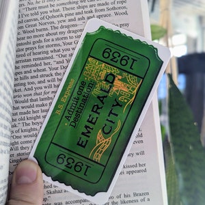 May include: A green and gold ticket with the words "Admit one. Destination: Emerald City" and the number "6261" printed on it. The ticket is partially covering a book with text about dragons.