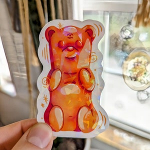 May include: A holographic sticker of a gummy bear with a third eye. The bear is orange and has a happy expression.