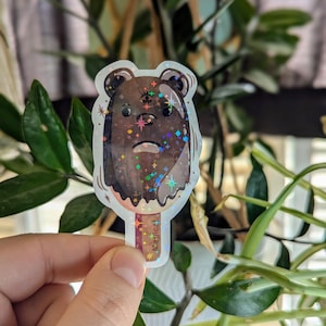 May include: A holographic sticker of a brown bear popsicle with a third eye and glitter. The sticker says "Magical" on the bottom.