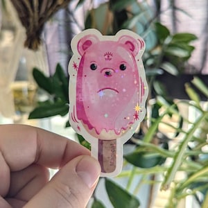 May include: A pink bear-shaped popsicle with a third eye and glitter. The popsicle is on a stick with the word "Magical" written on it.