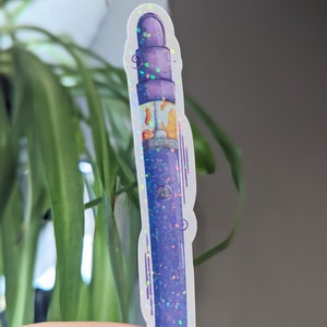 Peut inclure: Un autocollant de stylo vape en paillettes violettes et dorées. L'autocollant est holographique et a un contour blanc.