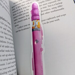 Peut inclure: Un marque-page rose et jaune avec une illustration stylisée d'un stylo et le texte "Ben's magic pens".
