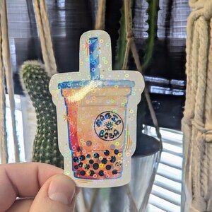 May include: A holographic sticker of a bubble tea cup with a straw and the text "Magic Boba" in a circle with a star and eye inside. The cup is filled with brown tea and tapioca pearls.