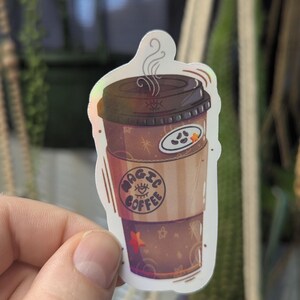 May include: A holographic sticker of a coffee cup with steam rising. The cup has a brown lid and a sleeve with the words "MAGIC COFFEE" and an eye design. A small star and other designs are visible.