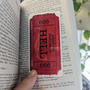 May include: A red ticket with the words "Hell" and "Destination" printed on it. The ticket has the number 666 printed on it twice. The ticket is sitting on top of an open book.