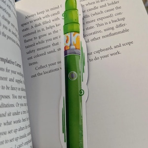 Peut inclure: Un marque-page vert et orange avec une illustration stylisée d'une cigarette électronique. Le marque-page porte le texte "Keep it real, ya'll".