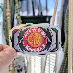 Könnte beinhalten: Aufkleber mit dem Power Rangers-Logo. Das Design umfasst einen roten Kreis mit einem Elefanten-Emblem, umgeben von den Worten "POWER RANGERS". Der Aufkleber hat einen schwarzen und silbernen Rand.