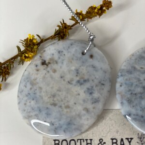 Op de afbeelding: Twee ronde, witte, grijze en zwarte gespikkelde keramische ornamenten met een zilveren touwtje aan elk. De ornamenten hangen aan een tak met gele bloemen. De ornamenten zijn gelabeld "Booth & Bay".