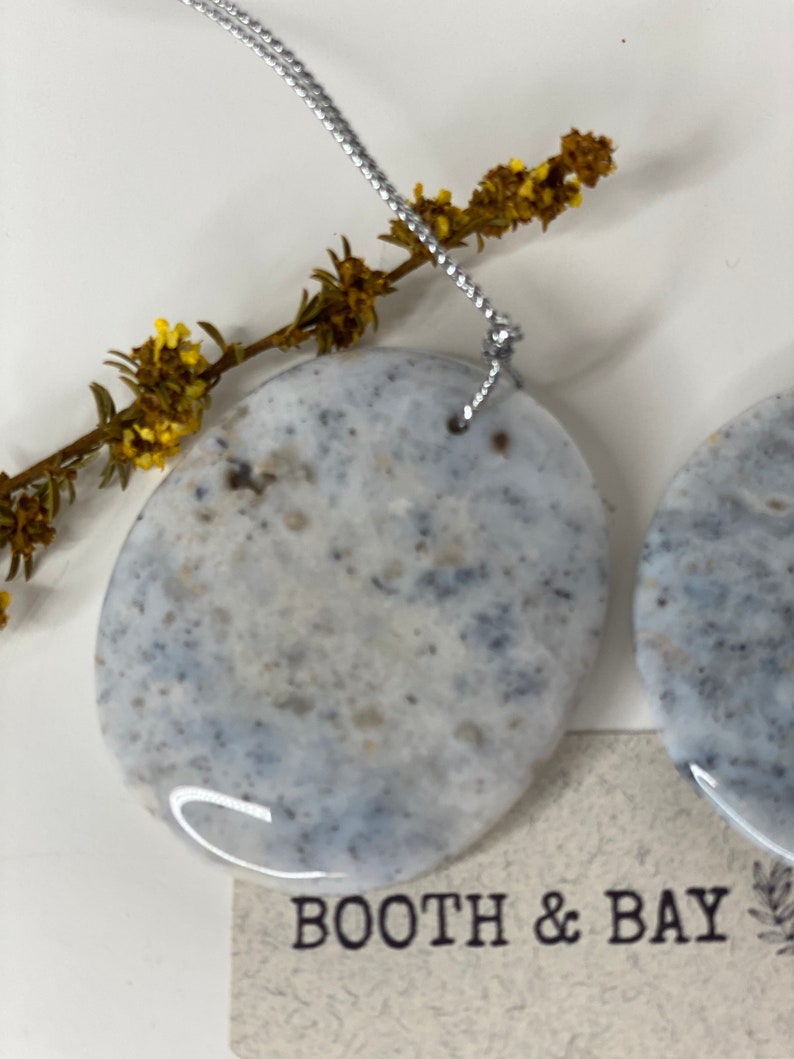 Op de afbeelding: Twee ronde, witte, grijze en zwarte gespikkelde keramische ornamenten met een zilveren touwtje aan elk. De ornamenten hangen aan een tak met gele bloemen. De ornamenten zijn gelabeld "Booth & Bay".