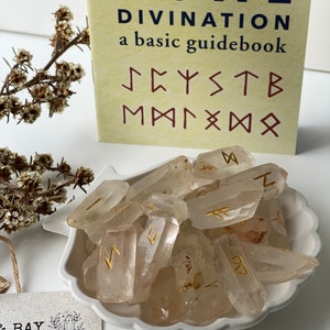 Könnte beinhalten: Ein Set aus klaren Quarzkristallen mit goldenen Runen, die darauf gemalt sind. Die Kristalle befinden sich in einer weißen Schale in Muschelform. Das Buch im Hintergrund trägt den Titel "Rune Divination: a basic guidebook".