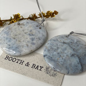 Op de afbeelding: Twee ronde, gepolijste stenen hangers in grijs en wit met een zilveren koord. De stenen hebben een marmerpatroon. De hangers hangen aan het koord en bevinden zich voor een witte achtergrond. De tekst "BOOTH & BAY" is zichtbaar op een klein bruin label.