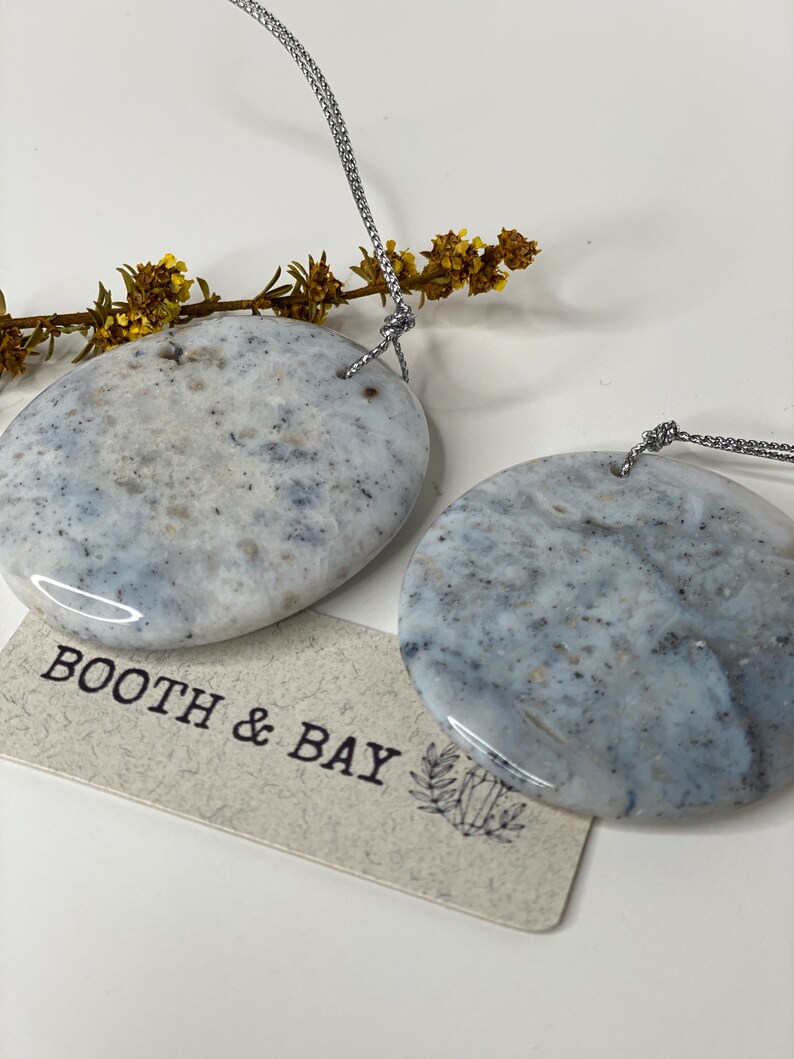 Op de afbeelding: Twee ronde, gepolijste stenen hangers in grijs en wit met een zilveren koord. De stenen hebben een marmerpatroon. De hangers hangen aan het koord en bevinden zich voor een witte achtergrond. De tekst "BOOTH & BAY" is zichtbaar op een klein bruin label.