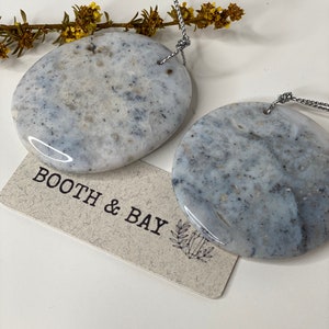 Op de afbeelding: Twee ronde, grijs en witte, marmeren stenen hangers met gaten voor een touwtje. De hangers hangen aan een zilveren touwtje. De hangers bevinden zich op een witte achtergrond met een klein takje gele bloemen. Een label met de tekst "Booth & Bay" bevindt zich in de linkerbenedenhoek van de afbeelding.