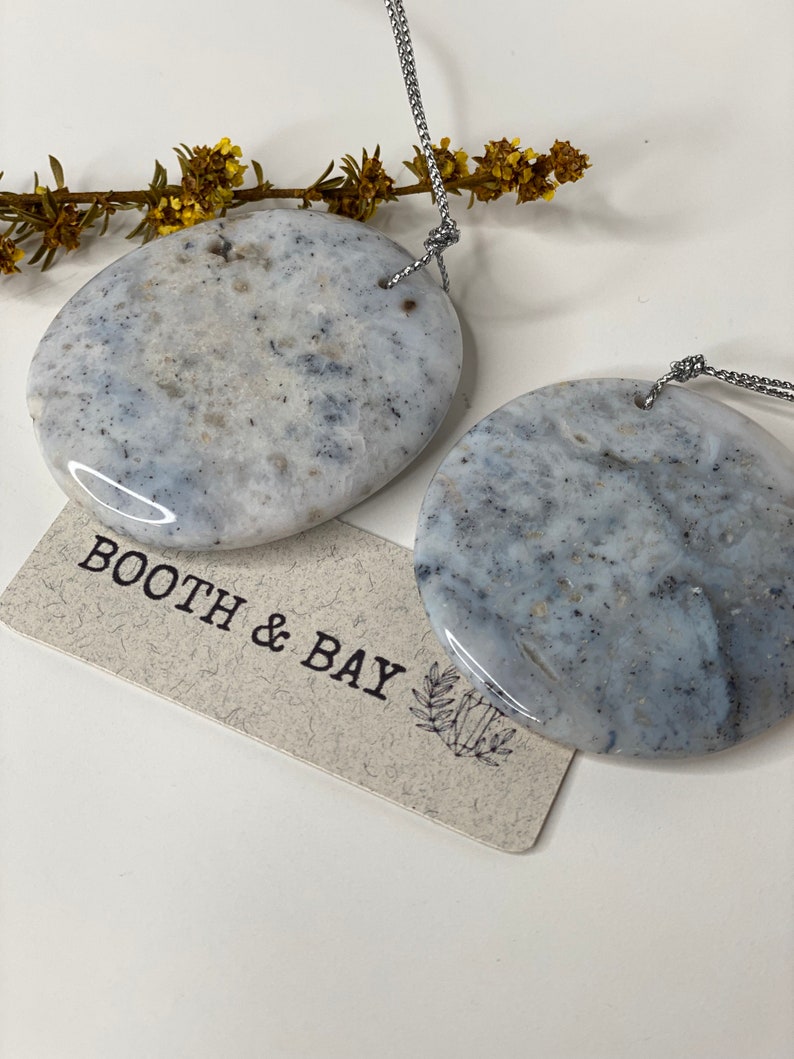 Op de afbeelding: Twee ronde, grijs en witte, marmeren stenen hangers met gaten voor een touwtje. De hangers hangen aan een zilveren touwtje. De hangers bevinden zich op een witte achtergrond met een klein takje gele bloemen. Een label met de tekst "Booth & Bay" bevindt zich in de linkerbenedenhoek van de afbeelding.