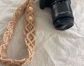 May include: A beige crocheted camera strap with wooden beads. The strap is attached to a black Canon camera with a lens.
