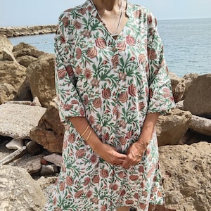 Könnte beinhalten: Ein weißes Baumwollkleid mit einem Blumendruck in Grün-, Rot- und Brauntönen. Das Kleid hat einen V-Ausschnitt, 3/4-Ärmel und einen Knopfleisten vorne. Das Kleid ist knielang und hat eine lockere Passform.