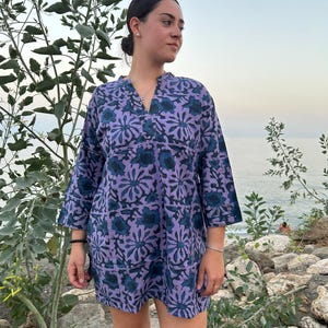 Op de afbeelding: Een vrouw draagt een korte mouw jurk met een paarse en blauwe bloemenprint. De jurk heeft een V-hals en een losse pasvorm.