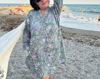 May include: A woman wearing a green floral print shirt dress with long sleeves and a button-up front. She is standing on a beach with the ocean in the background.