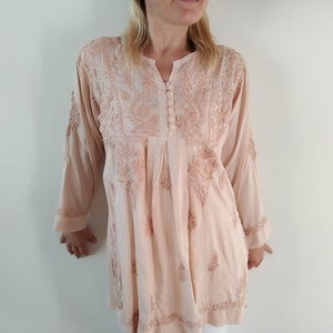 Op de afbeelding: Een lichtroze, langarmige tuniek met ingewikkelde bloemenborduursels. De tuniek heeft een V-hals en een knoopsluiting aan de hals.