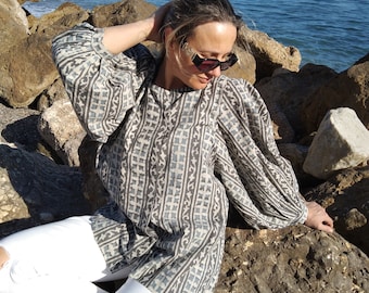 May include: A woman wearing a gray and white patterned blouse with long sleeves and white jeans sits on rocks by the ocean.