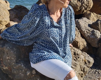 May include: A woman wearing a blue and white patterned blouse with a white v-neck and long sleeves. She is wearing white jeans with a distressed knee and is sitting on a large rock.