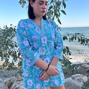 Op de afbeelding: Een vrouw draagt een blauwe tuniek met witte bloemenprint, een V-hals en 3/4 mouwen. De tuniek heeft een witte rand rond de hals en mouwen. De vrouw staat voor een waterpartij.