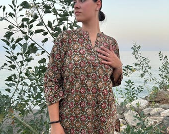 May include: A woman wearing a green and red floral print tunic dress with a V-neckline. The dress has 3/4 sleeves and a tie at the side. She is standing on a beach with rocks and water in the background.