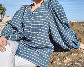 May include: A woman wearing a blue and white plaid shirt with long sleeves and a relaxed fit. She is sitting on a rock with her legs crossed and wearing white jeans.