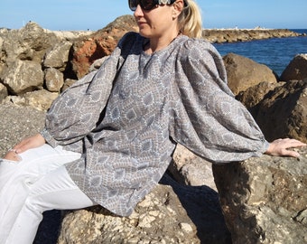 May include: A woman wearing sunglasses sits on a rocky shore wearing a gray and white patterned blouse with puffed sleeves. The woman is wearing white jeans and has her hair pulled back.