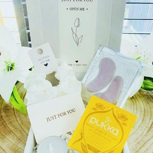May include: A gift box with the text "JUST FOR YOU" and "OPEN ME". Inside are a white scrunchie, a tea bag, a small candle, and two pink facial tools. The box also contains a card that says "Have a nice day".