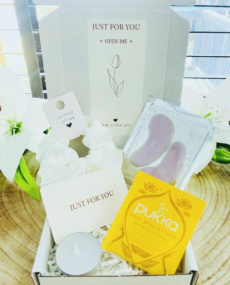 May include: A gift box with the text "JUST FOR YOU" and "OPEN ME". Inside are a white scrunchie, a tea bag, a small candle, and two pink facial tools. The box also contains a card that says "Have a nice day".