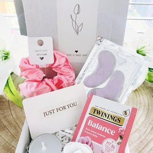 May include: A gift box containing a pink scrunchie, eye patches, a tea bag, a candle, and a decorative flower. The box includes a card that says "JUST FOR YOU" and "+ OPEN ME +" with a tulip illustration. A small tag reads "Handmade with love."