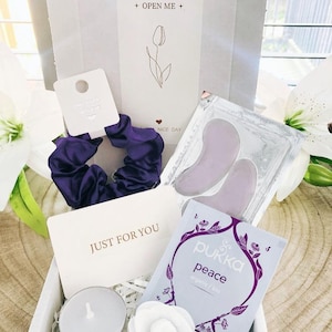 May include: A gift box containing a purple scrunchie, a tea bag, eye patches, a candle, and a white paper flower. The box includes a card that reads "JUST FOR YOU" and "OPEN ME". The tea bag is labeled "Pukka peace organic/bio".