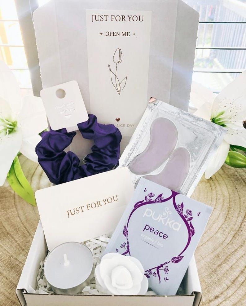 May include: A gift box containing a purple scrunchie, a tea bag, eye patches, a candle, and a white paper flower. The box includes a card that reads "JUST FOR YOU" and "OPEN ME". The tea bag is labeled "Pukka peace organic/bio".