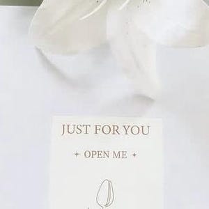 May include: A white lily flower with green leaves and a card that reads "JUST FOR YOU + OPEN ME +" with a line drawing of a tulip. The image is set against a white background.