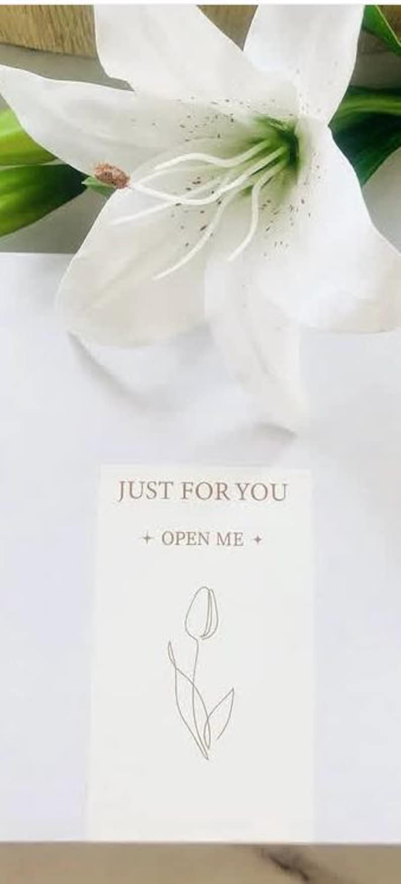 May include: A white lily flower with green leaves and a card that reads "JUST FOR YOU + OPEN ME +" with a line drawing of a tulip. The image is set against a white background.