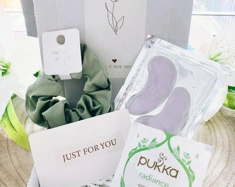 May include: A gift box containing a variety of self-care items. Includes a green scrunchie, eye patches, a tea bag, a white rose-shaped soap, and a small candle. The box has a card that says "JUST FOR YOU" and "+ OPEN ME +."