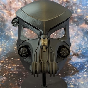 May include: A black plastic Predator mask with a detailed design, including a visor, a mouth piece, and a series of circular details on the sides. The mask is displayed on a black stand.