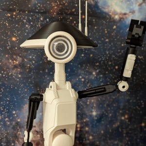 May include: A white and black articulated robot with a round, black and white head and a single, large, round eye. The robot is posed with one arm raised in a wave. The robot is set against a dark blue background with white stars.