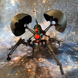 Könnte beinhalten: Ein schwarzes Robotermodell mit zwei großen, halbkreisförmigen Strukturen und mehreren gelenkigen Beinen. Das Modell hat rote kreisförmige Akzente und eine zentrale Antenne. Der Hintergrund ist eine Sternengalaxie.